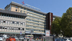 Hospital building facade with large signage reading OSPEDALE GIOVANNI BOSSO, photographed outdoors under a blue sky with trees and parked cars in the foreground.