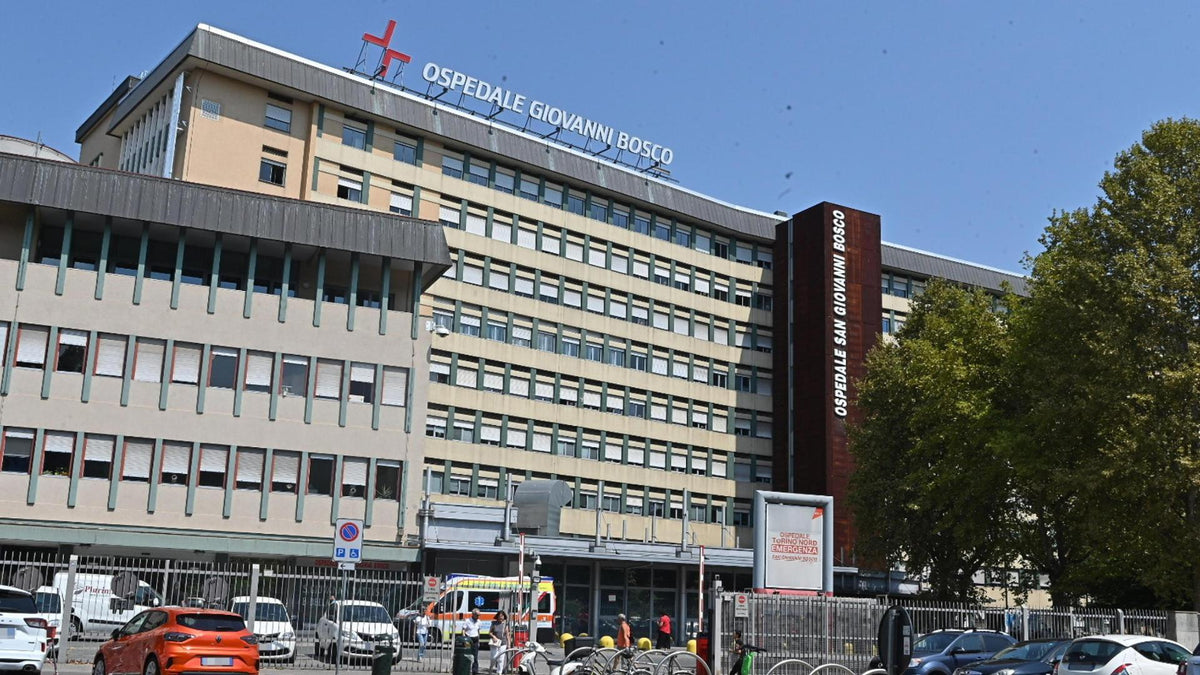 Hospital building facade with large signage reading OSPEDALE GIOVANNI BOSSO, photographed outdoors under a blue sky with trees and parked cars in the foreground.